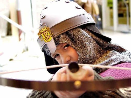 Ein Kind in mittelalterlicher Verkleidung mit Kettenhaube und Helm zielt mit einer Armbrust direkt in die Kamera.