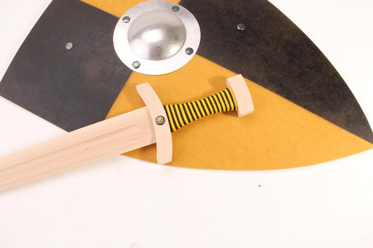 Nahaufnahme eines schwarz-gelb geviertelten Spielzeugschildes mit silbernem Schildbuckel. Darauf liegt ein Holzschwert mit gelb-schwarz umwickeltem Griff und Parierstange aus Holz.