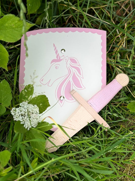 Holzschild mit rosa Einhorn-Motiv und passendem Holzschwert mit hellrosa Griff liegen im grünen Gras. Das Schild hat einen rosa Rand mit Wellenmuster, das Schwert lehnt dekorativ daran. Im Vordergrund befindet sich eine blühende Wildblume.