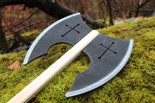 Doppelseitige Spielzeug-Axt aus Holz mit schwarzem Axtkopf und eingravierten Kreuzen liegt auf moosbedecktem Boden im Wald.