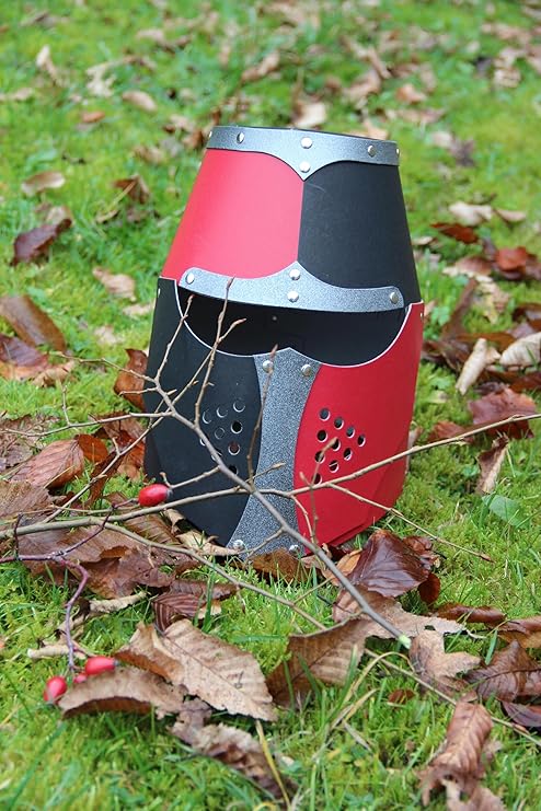 Normannenhelm für Kinder aus bemaltem Material in Schwarz, Rot und Silber mit Nieten und Lüftungslöchern. Der Helm steht auf grünem Gras, umgeben von braunen Herbstblättern und einem Zweig mit roten Beeren.