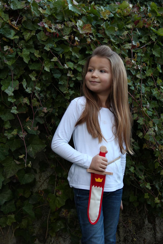 Ein Kind mit langen, hellbraunen Haaren steht draußen vor einer Efeuwand. Es trägt ein weißes Langarmshirt und eine blaue Jeans. In der rechten Hand hält es ein Holzspielzeug-Schwert in einer weißen Scheide mit rotem Rand und gelbem Kronenmotiv.