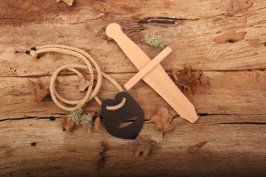 Holzschwert und Schwertträger „Kuno“ mit brauner Trageschnur und dunklem Halter aus Recyclingleder liegen auf rustikalem Holzuntergrund, umgeben von Laub und Moos.