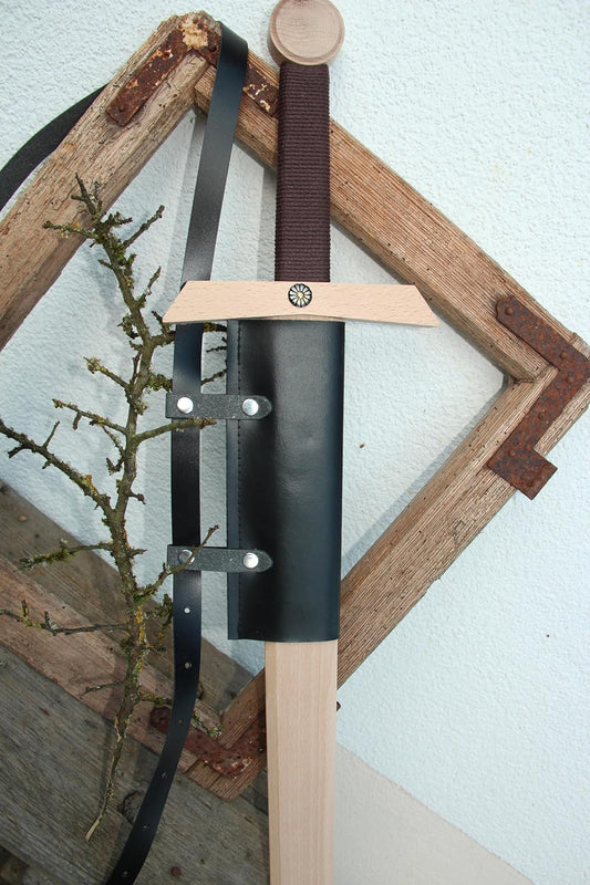 Holzschwert mit braun umwickeltem Griff in schwarzer Rückenscheide aus Leder, an einem rustikalen Holzrahmen lehnend.