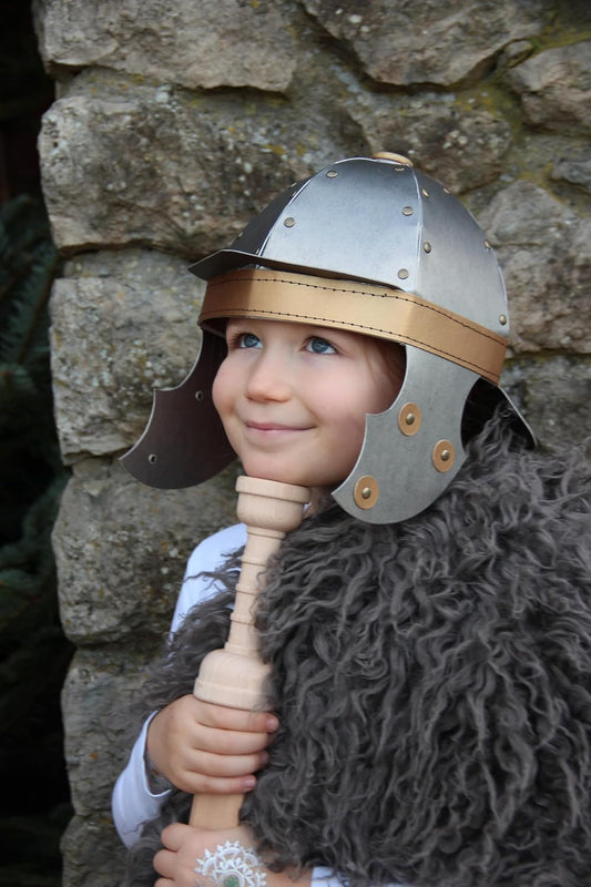 Kind mit römischem Helm und grauem Fellumhang steht vor einer Steinmauer und hält ein naturfarbenes Holzschwert. Das Kind lächelt und blickt nach oben.