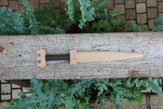 Holzspielzeug-Dolch mit grünem Griffband und gezacktem Knauf, liegend auf einem rustikalen Holzbalken, umgeben von grünen Pflanzen im Freien.