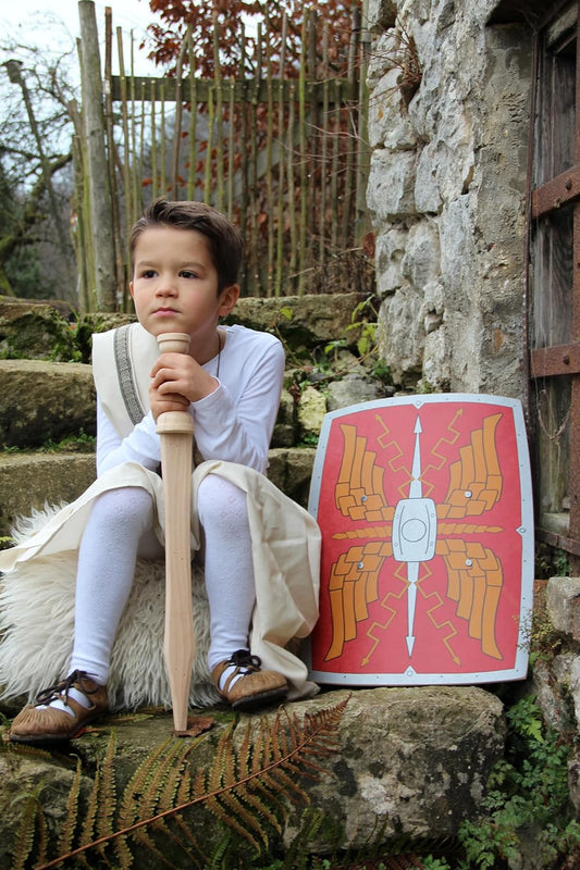 Kind in römischer Tunika sitzt mit einem Holzschwert auf einer Steintreppe. Rechts daneben steht ein rechteckiger, rot bemalter Römer-Schild mit Flügelmotiv und weißem Zierbeschlag.