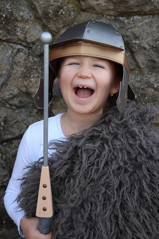 Ein Kind steht vor einer Felswand, trägt einen silbernen römischen Helm, einen dicken grauen Schaffellumhang und hält lachend einen Holzspeer mit grauer Spitze in der Hand.