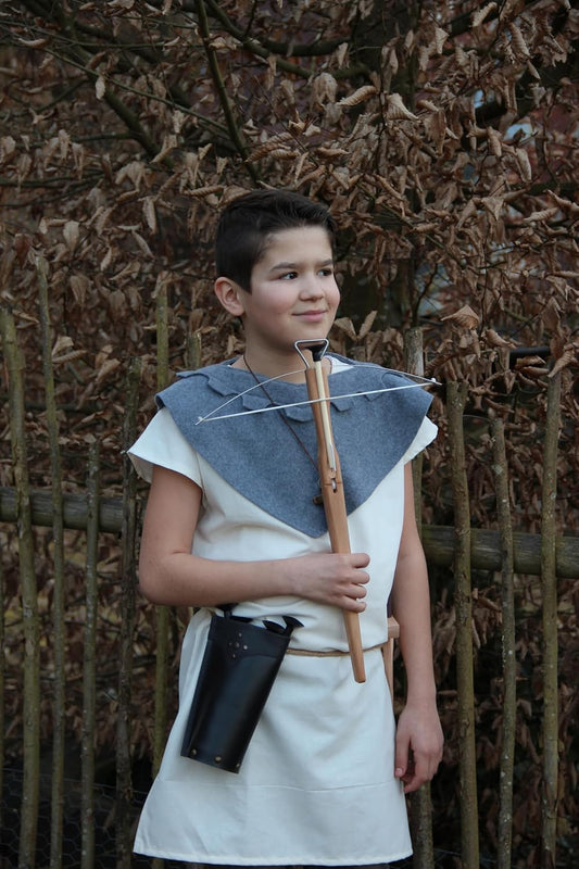 Junge in historischer Gewandung mit weißem Gewand, blauem Schultertuch und schwarzem Köcher, hält eine hölzerne Armbrust in der Hand, im Hintergrund ein Holzzaun und braune Laubäste.