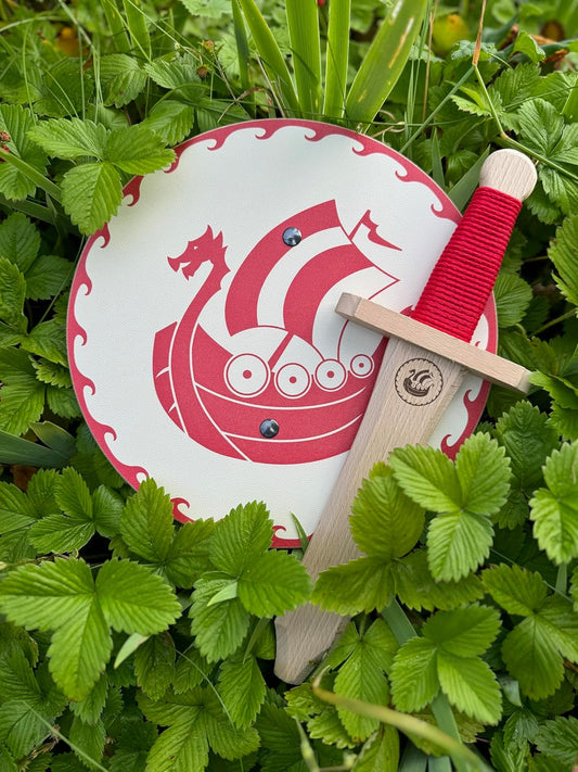 Kinderschild mit rotem Drachenbootmotiv und Wellenrand sowie passendes Holzschwert mit roter Griffwicklung und Wikingergravur, arrangiert im grünen Erdbeerlaub.