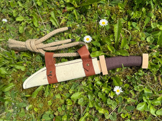 Holzspielzeugmesser in heller Filzscheide mit braunen Lederriemen und Kordel liegt auf grünem Gras mit neben Gänseblümchen.