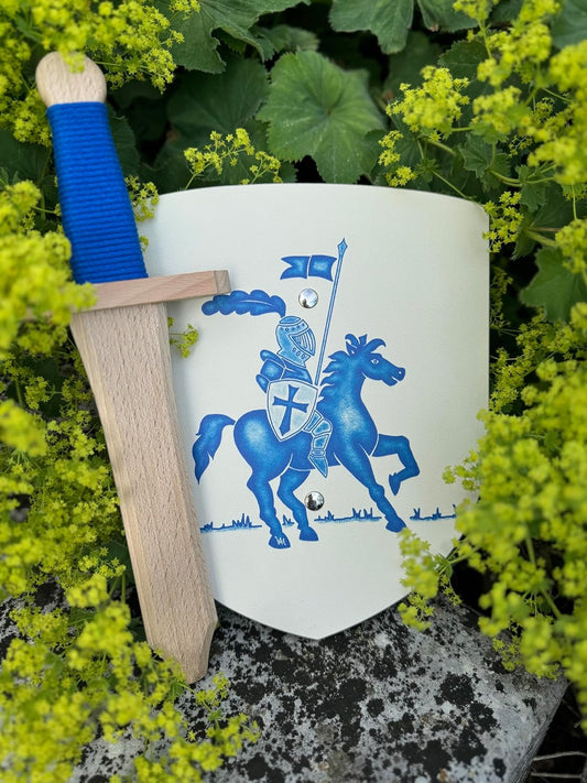 Holzschwert mit blauem Griff liegt an einem weißen Holzschild mit blauem Rittermotiv, umgeben von grünen Blättern und gelb blühenden Pflanzen.