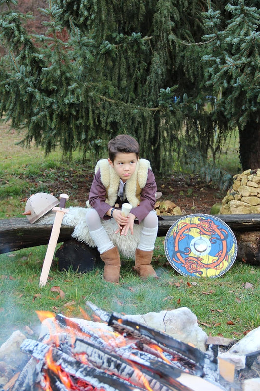 Ein Junge in historischer Verkleidung sitzt auf einem mit Fell bedeckten Holzstamm vor einem Lagerfeuer. Neben ihm liegen ein Holzschwert, ein Helm und ein rundes Schild mit buntem Muster. Im Hintergrund steht eine große Tanne.