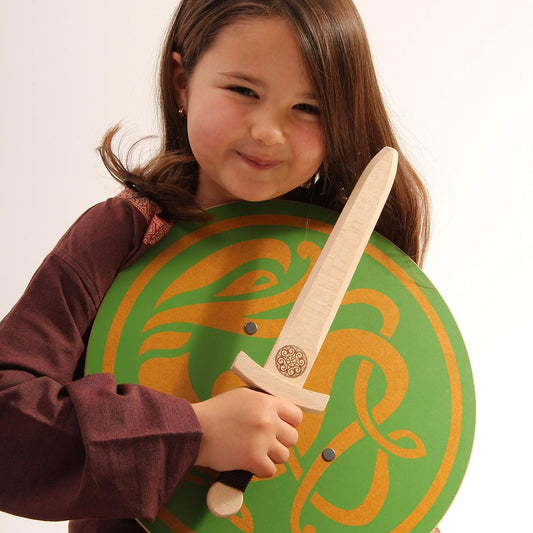 Kind in braunem Gewand hält lachend ein Holzschwert und einen grünen Rundschild mit goldener keltischer Tierverzierung. Das Schwert zeigt ein keltisches Muster an der Parierstange.