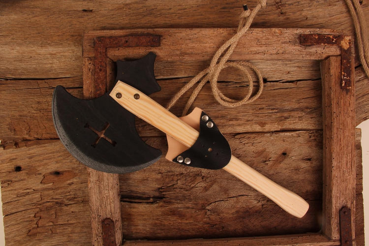 Spielzeug-Holzaxt mit naturbelassenem Holzgriff und schwarzem Axtkopf, der ein eingraviertes Kreuzsymbol zeigt. Die Axt steckt in einer schwarzen Gürtelschlaufe aus Lederimitat. Präsentiert auf rustikalem Holzuntergrund mit grobem Seil.