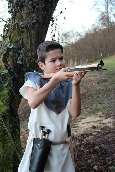 Junge in historischer Gewandung mit weißem Gewand, blauem Schultertuch und schwarzem Köcher zielt mit einer hölzernen Armbrust, im Hintergrund ein mit Efeu bewachsener Baum und eine waldige Landschaft.