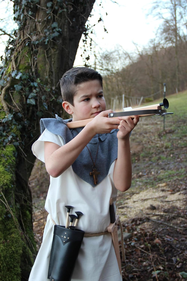 Junge in historischer Gewandung mit weißem Gewand, blauem Schultertuch und schwarzem Köcher zielt mit einer hölzernen Armbrust, im Hintergrund ein mit Efeu bewachsener Baum und eine waldige Landschaft.