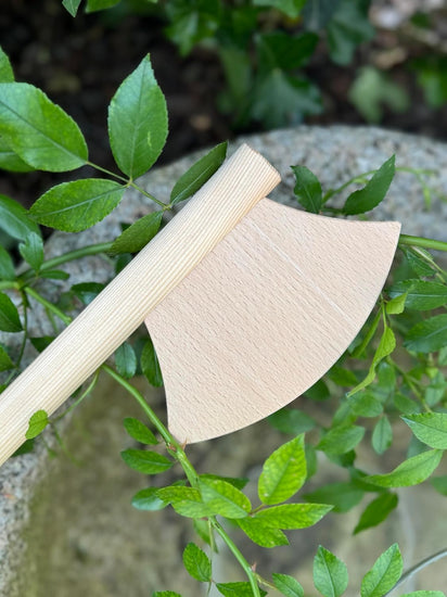 Holzspielzeug-Axt mit naturbelassenem Stiel und hellem Axtkopf, liegend auf grünem Blattwerk vor einem Stein.