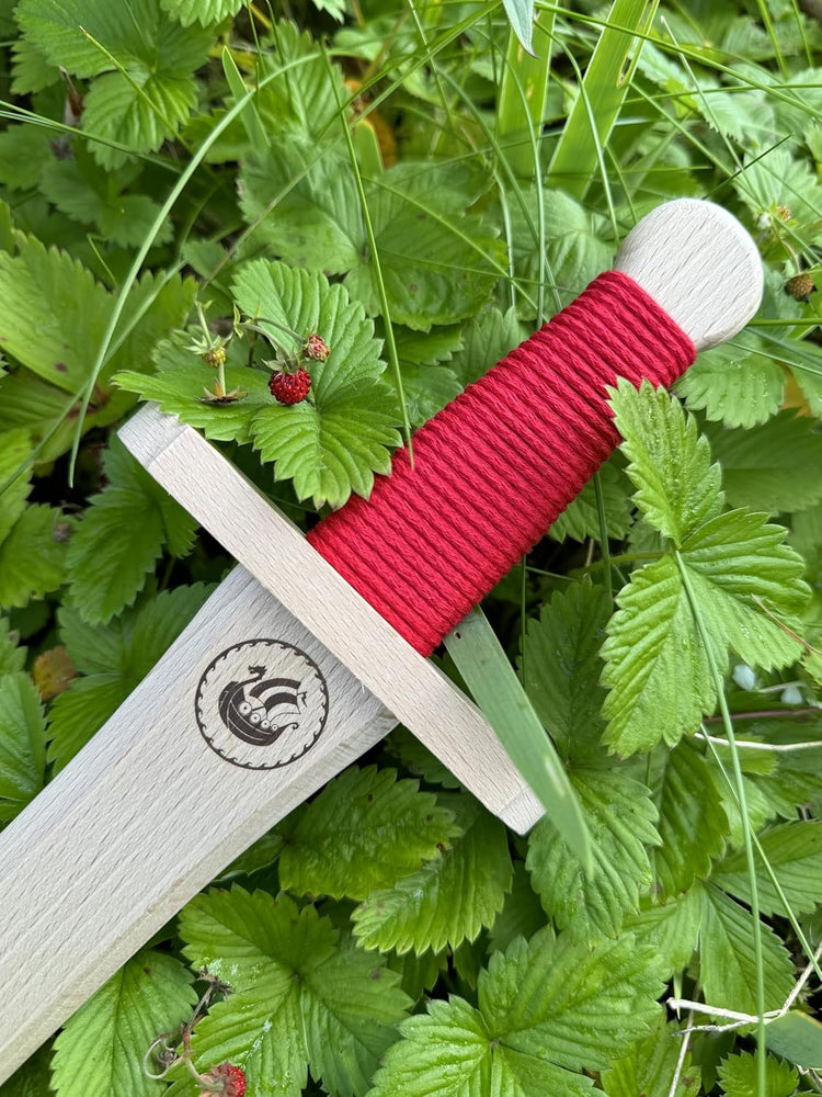 Detailaufnahme eines Holzschwerts mit roter Griffwicklung und eingraviertem Drachenboot-Motiv, eingebettet in grüne Erdbeerblätter mit kleinen Walderdbeeren.