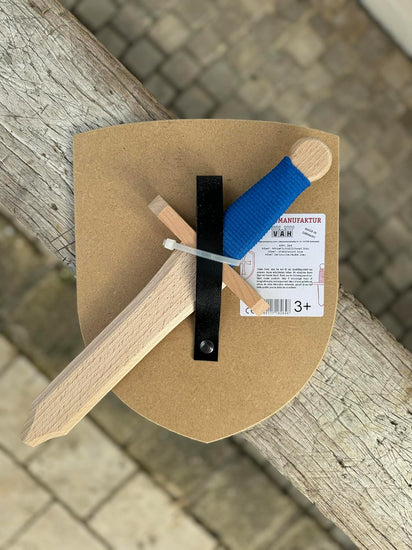 Rückseite des Spielsets der Spielzeugmanufaktur VAH, Artikelnummer 264: Naturfarbener Holzschild mit schwarzer Halterung und befestigtem Holzschwert mit blauer Griffwicklung. Auf dem Schild ist ein Etikett mit Produktinformationen und Altersempfehlung ab 3 Jahren angebracht.