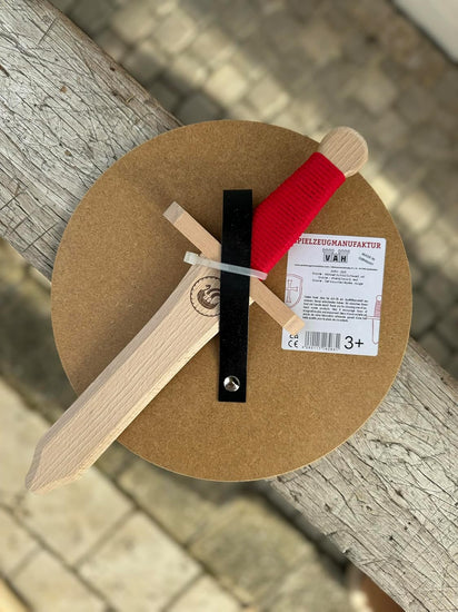 Rückseite eines Kinderschilds mit schwarzer Halteschlaufe und befestigtem Holzschwert mit roter Griffwicklung und Wikingergravur. Rechts ein Etikett mit VAH-Logo und Altersangabe 3+.