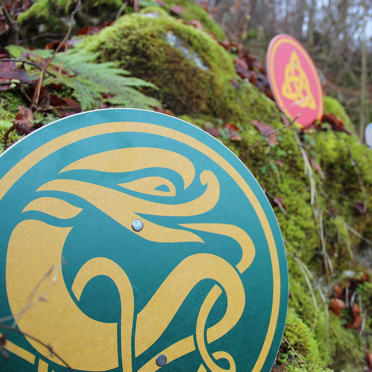 
Zwei bemalte Rundschilde im Moos und Laub eines Waldbodens. Vorderer Schild grün mit goldener keltischer Tierfigur, hinterer Schild rot mit gelbem Knotenmotiv, teils unscharf im Hintergrund.
