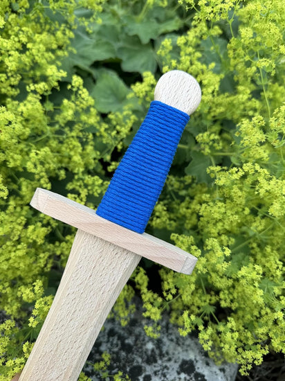 Nahaufnahme eines Holzschwertes der Spielzeugmanufaktur VAH mit blau umwickeltem Griff, einfachem Handschutz und runder Knaufkappe, im Hintergrund grüne Pflanzen mit gelben Blüten.