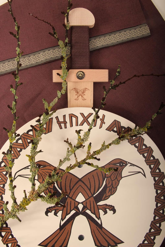 Dekoratives Arrangement mit Holzschwert, rundem Schild mit Runenschrift und zwei stilisierten Raben, violett-braunem Stoff mit Zierborte und einem Zweig mit Flechten. Auf dem Schwertgriff ist ein Adlermotiv eingraviert.