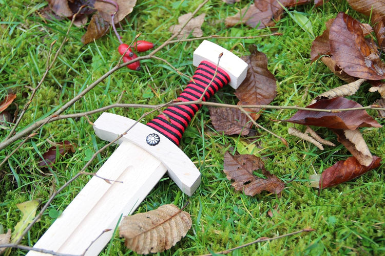 Holzschwert mit naturfarbener Klinge und rot-schwarz umwickeltem Griff liegt auf moosigem Boden mit Herbstlaub, Zweigen und Hagebutten.