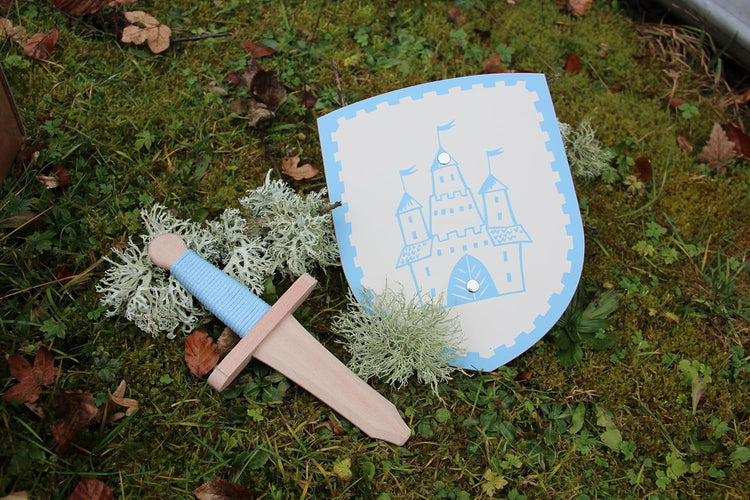 Kinderspielzeug-Set bestehend aus einem Holzschwert mit hellblauem Griff und einem passenden Schild mit hellblauem Burgmotiv. Das Set liegt auf moosbedecktem Boden, dekoriert mit hellgrünem Islandmoos und herbstlichen Blättern.